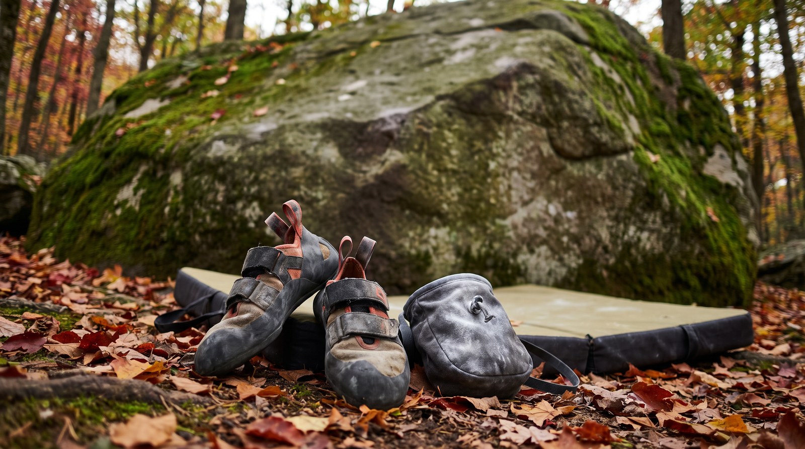 Kletterschuhe, ein Chalkbag und ein Crashpad liegen am Fuß eines Felsblocks im herbstlichen Wald