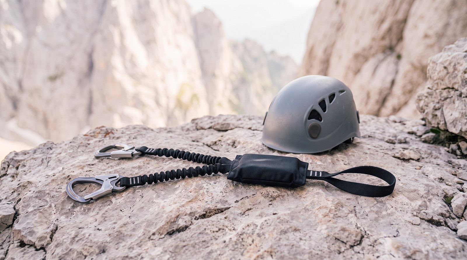 Ein Klettersteigset mit Bandfalldämpfer und zwei Karabinern sowie ein Kletterhelm liegen auf einem Felsabsatz, dahinter helle Felswände