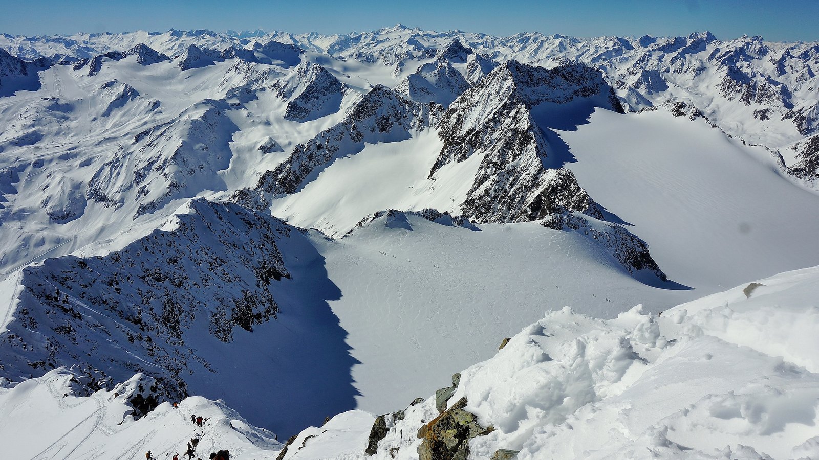 Verschneite Gipfel und ein Gletscherbecken im Skigebiet Stubaier Gletscher, im Vordergrund Tourengeher auf einem Grat