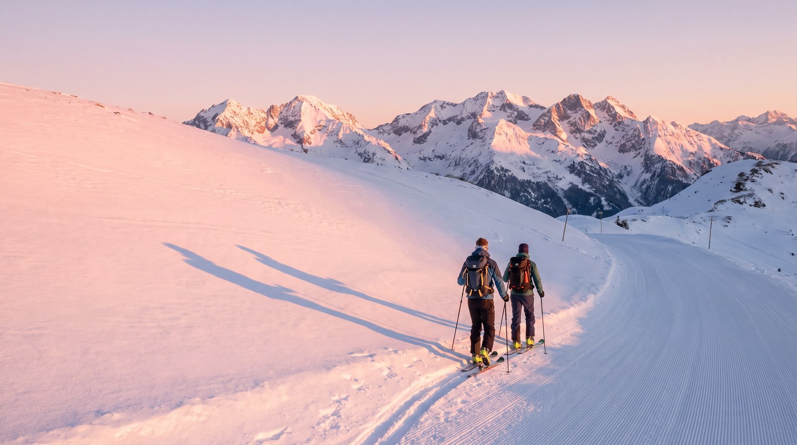 Zwei Skitourengeher steigen im ersten Morgenlicht am Rand einer präparierten Piste mit Fellen bergauf